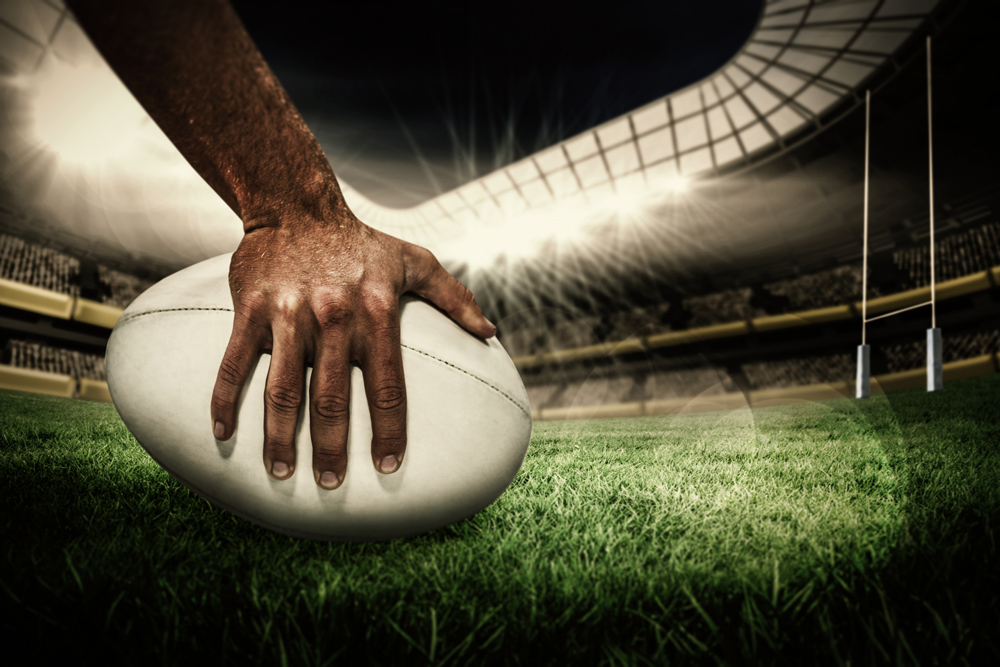 A rugby ball on the ground with a rugby players hand on it in a lit up stadium