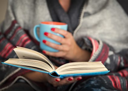 A woman snuggled in a blanket with a warm drink reading a book