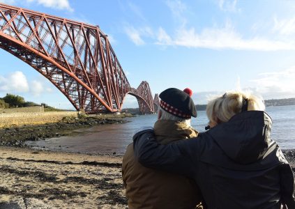 A couple sitting on the shores of the Forth Road Bridge looking up at the Forth Road Bridge