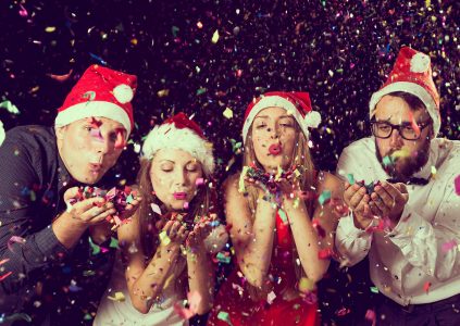 Two men and two women in Santa hats blowing Christmas confetti